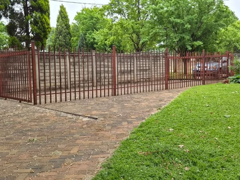 A rear garden area featuring a paved driveway, a lawn, and secure steel palisade fencing with a precast concrete boundary wall.