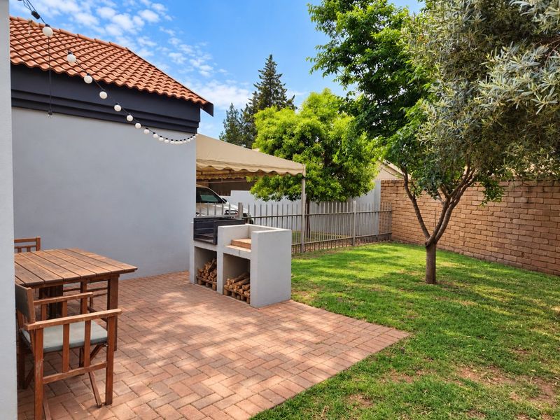 A well-maintained outdoor patio area featuring a built-in braai, paved flooring, and a private garden space with established trees and boundary walling.