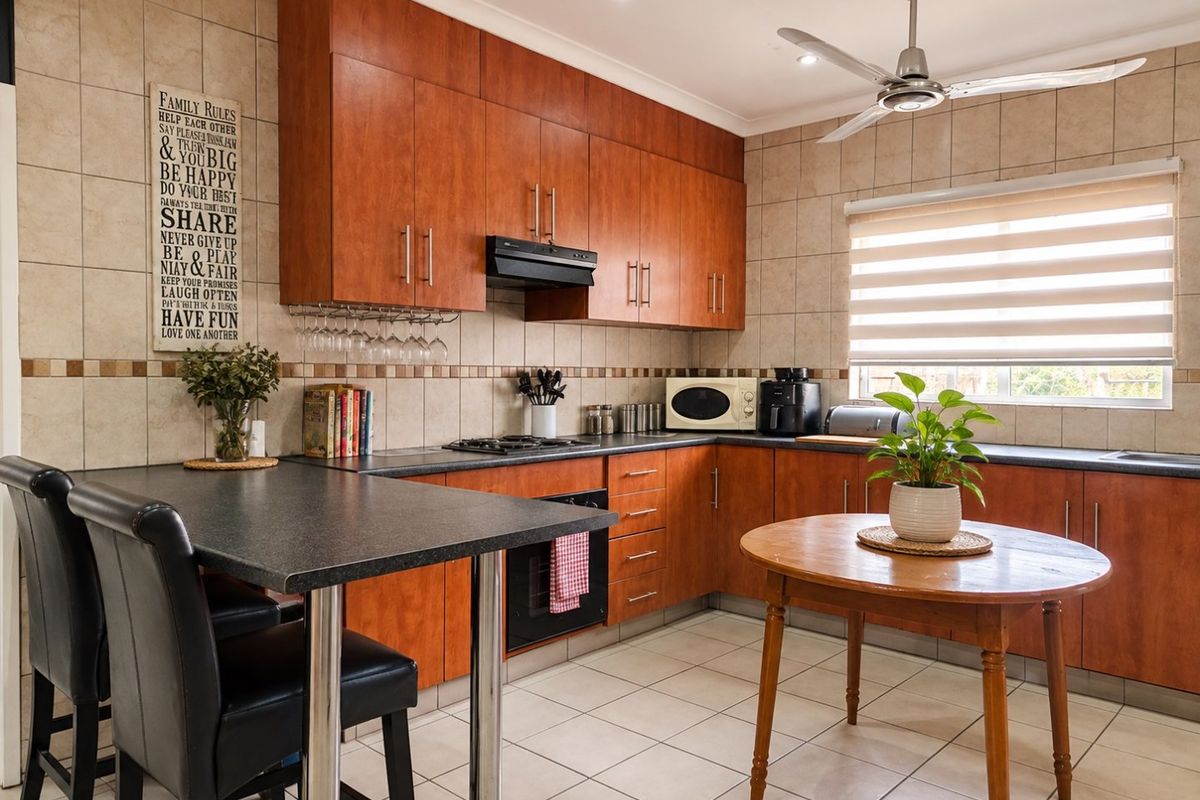 A functional kitchen featuring warm-toned wooden cabinetry, dark laminate countertops, built-in oven and hob, tiled backsplash, and a ceiling fan for ventilation.