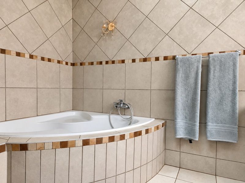 A bathroom corner bathtub featuring neutral ceramic tiling with a decorative border, chrome fixtures, and a wall-mounted light.