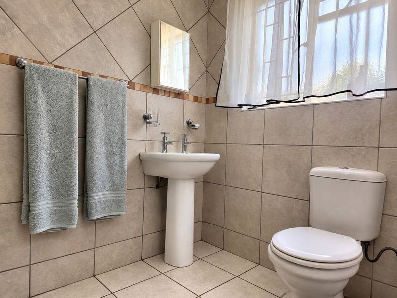A functional bathroom featuring a pedestal basin, toilet, and neutral wall and floor tiling with a window for natural light.