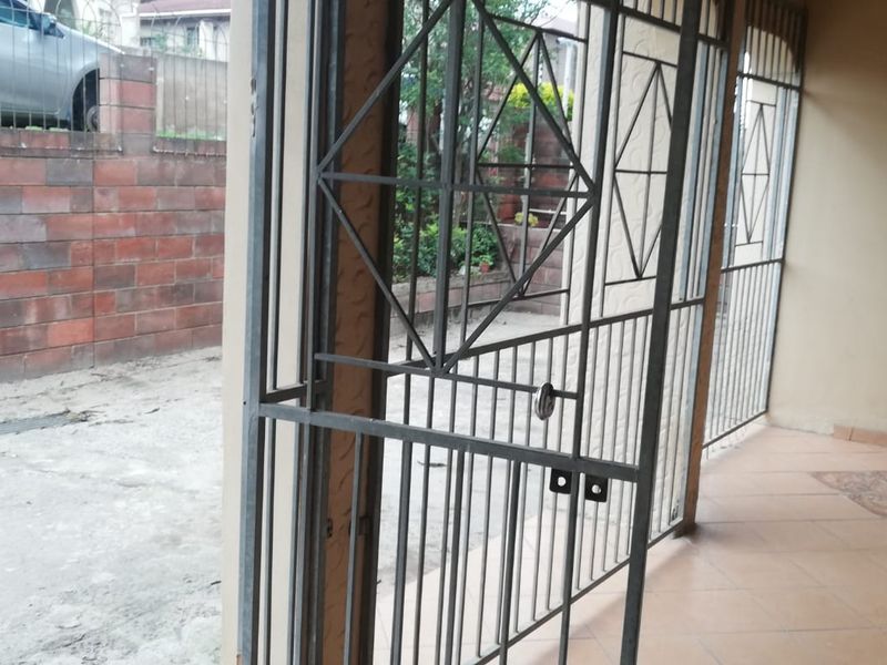 A covered patio area featuring tiled flooring and a security gate, overlooking a paved outdoor space with a brick retaining wall.