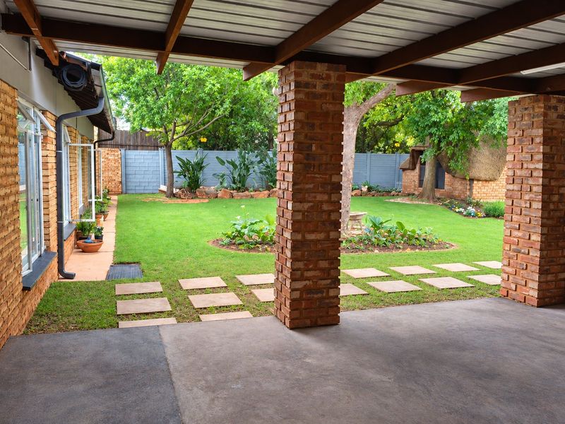 A covered patio area featuring exposed brick pillars and timber beams, overlooking a well-maintained garden with established trees and lawn.