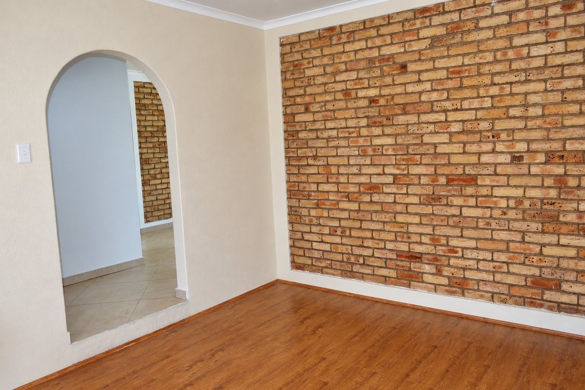An empty living room featuring a prominent exposed brick accent wall, warm laminate flooring, and a distinctive arched doorway leading to an adjacent space.