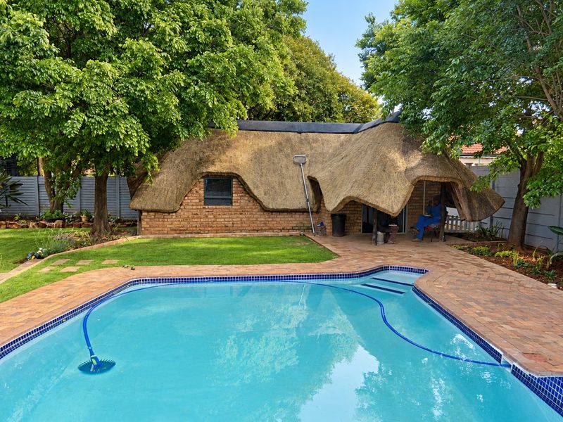 A well-maintained outdoor area featuring a swimming pool, paved patio, and a charming thatched-roof structure set within a landscaped garden.