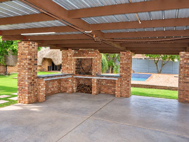 Covered outdoor patio featuring a built-in brick braai, concrete flooring, and exposed timber beams, overlooking the garden and swimming pool area.
