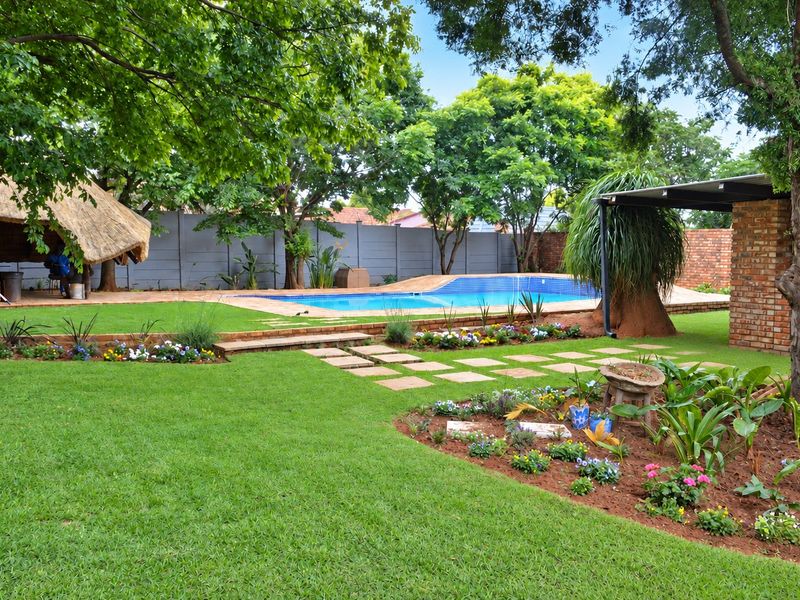 A well-maintained backyard featuring a swimming pool, a traditional thatched lapa, and a landscaped garden with mature trees and paved pathways.