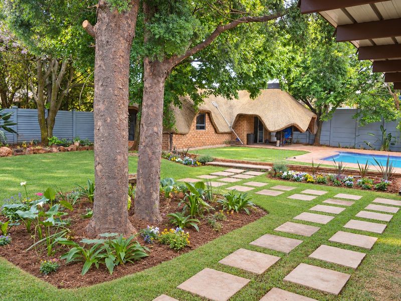 A well-maintained rear garden featuring a swimming pool, paved walkways, mature trees, and a charming thatched-roof outbuilding.
