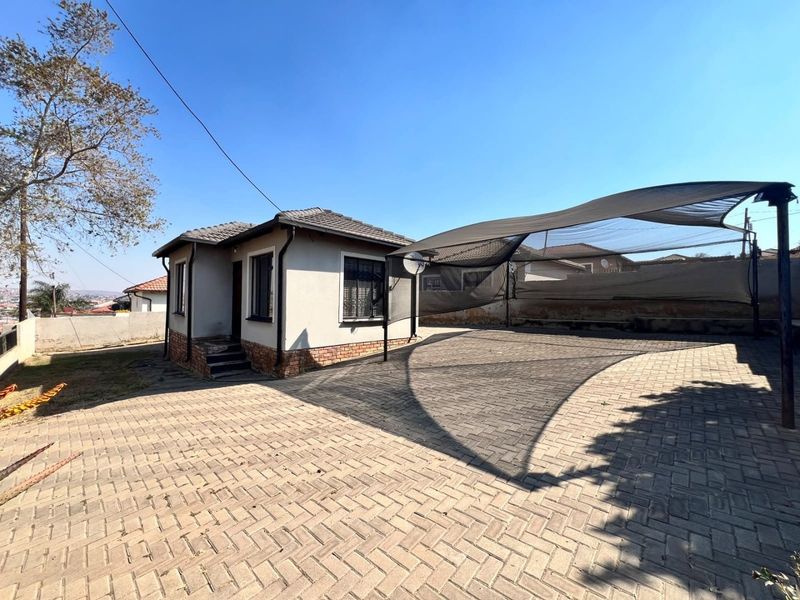 Front exterior view of the house featuring a large paved driveway, shade cloth carport, and tiled roof with security bars on windows.