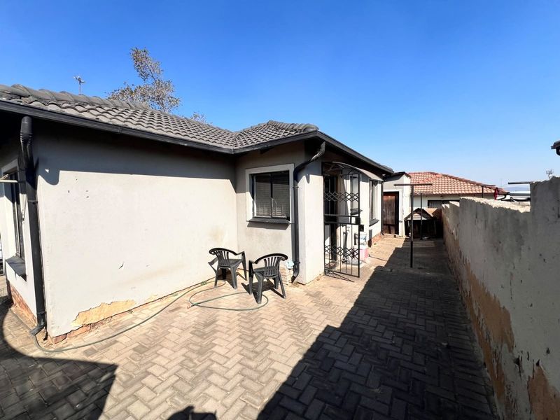 Rear exterior view of the house featuring a paved patio area, security gate, and boundary wall with visible signs of wear and peeling paint.