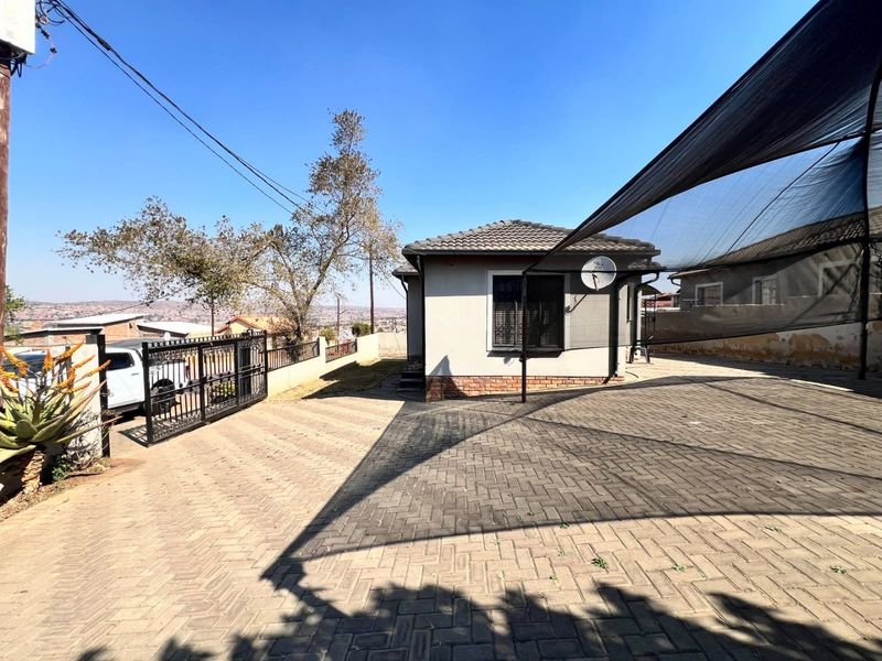 Front exterior view of the property featuring a paved driveway, shade cloth carport, and secure metal gate access.