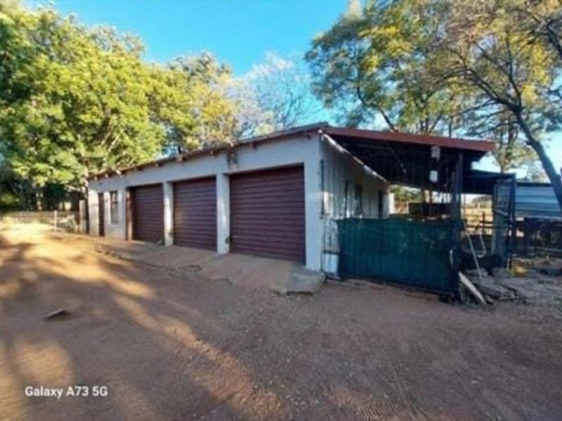 A triple garage structure with roll-up doors and an attached side carport, situated on a dirt driveway in a rural farm setting.