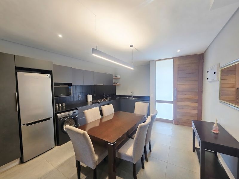 Modern open-plan kitchen and dining area featuring sleek dark cabinetry, integrated appliances, tiled flooring, and a contemporary timber entrance door.