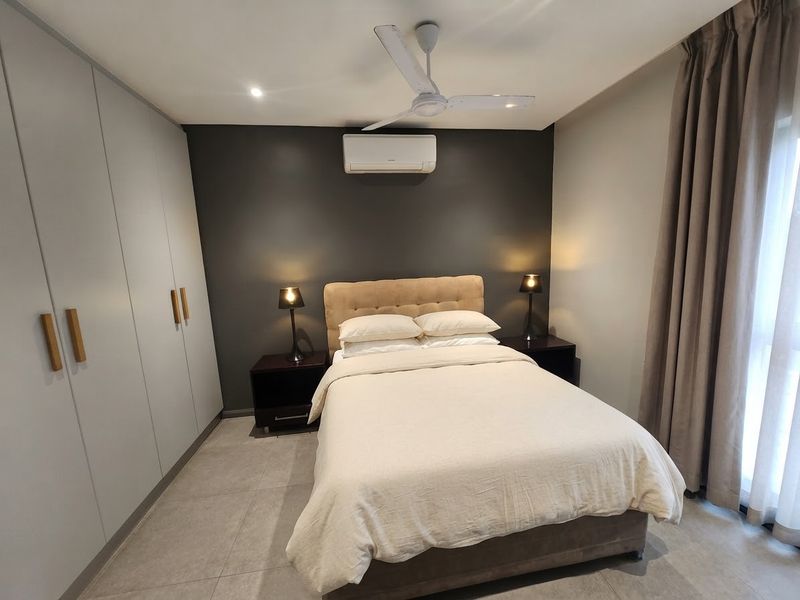 A well-maintained bedroom featuring neutral tiled flooring, built-in cupboards, a ceiling fan, and a wall-mounted air conditioning unit.