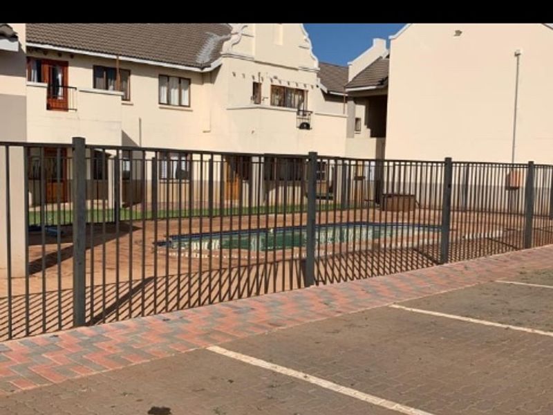 Exterior view of an apartment complex featuring a communal swimming pool enclosed by metal fencing and adjacent paved parking area.