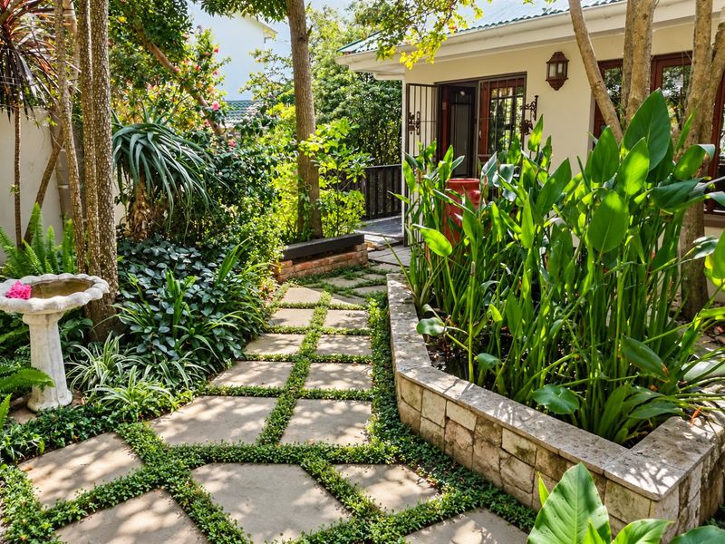 A lush, well-maintained garden featuring a stone-paved pathway, a raised stone planter with aquatic plants, and mature trees leading to the home entrance.