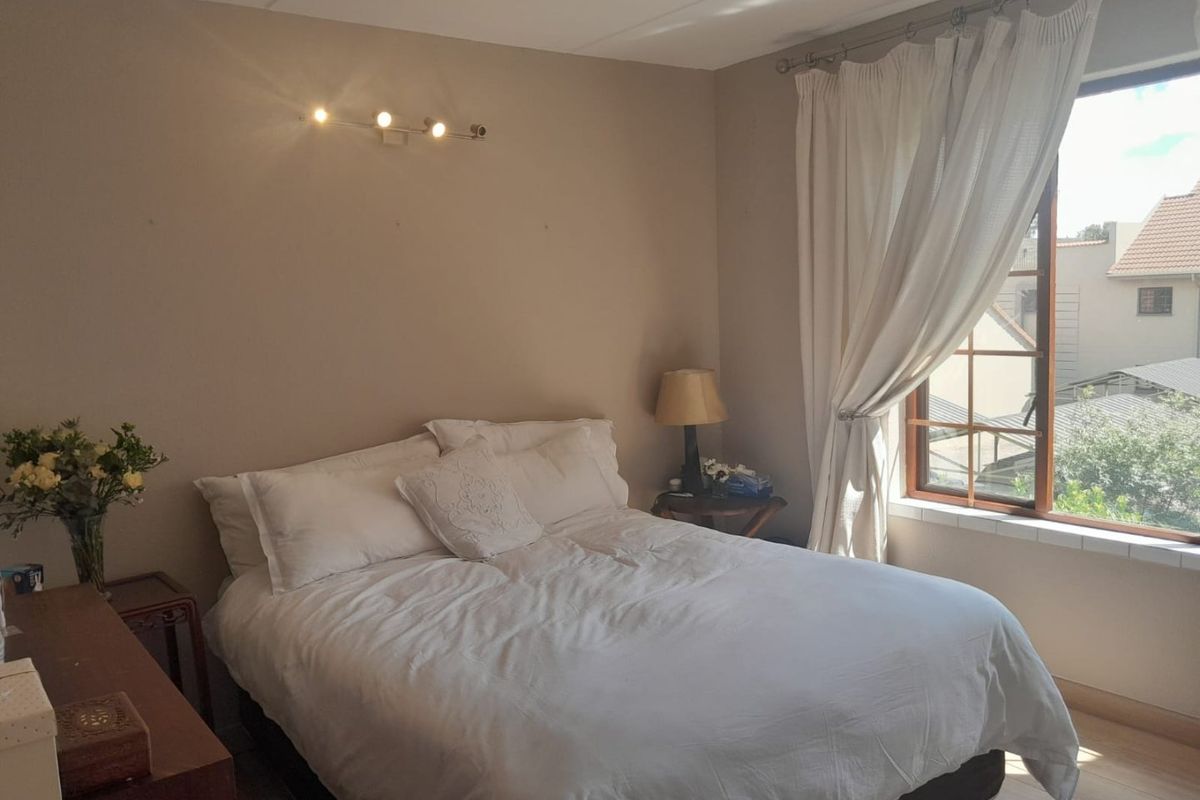 A cozy bedroom featuring a large window with natural light, neutral wall tones, and a wall-mounted spotlight fixture.