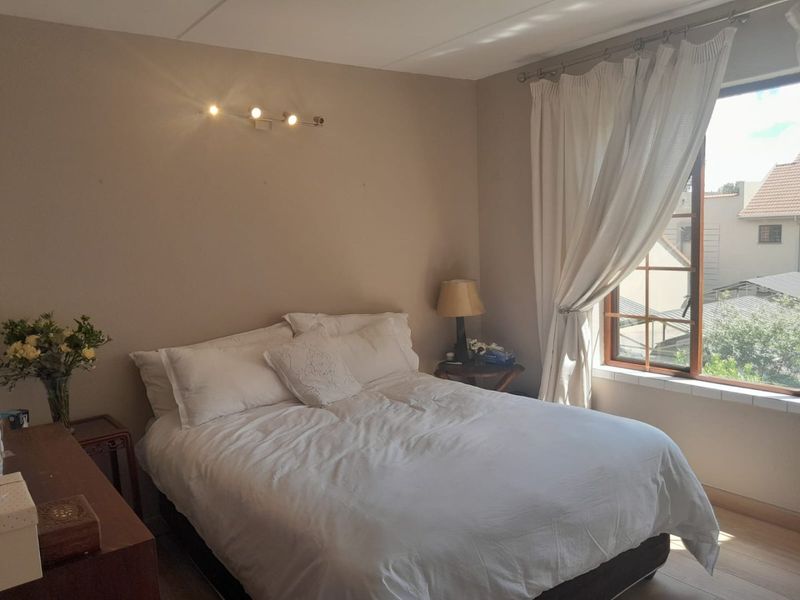A cozy bedroom featuring a large window with natural light, neutral wall tones, and a wall-mounted spotlight fixture.