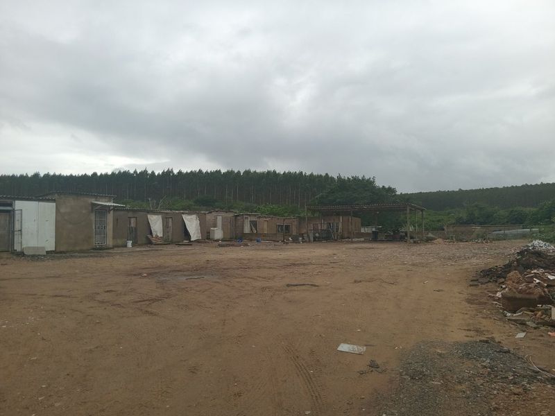 An expansive outdoor area featuring multiple basic outbuildings and a covered structure, set against a backdrop of trees on a large, unpaved plot.