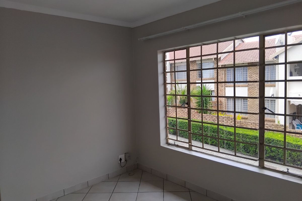 A bright, empty bedroom featuring neutral wall tones, tiled flooring, and a large window fitted with security bars overlooking the complex grounds.
