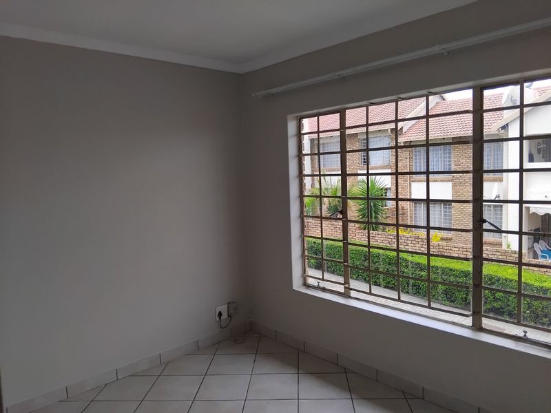 A bright, empty bedroom featuring neutral wall tones, tiled flooring, and a large window fitted with security bars overlooking the complex grounds.