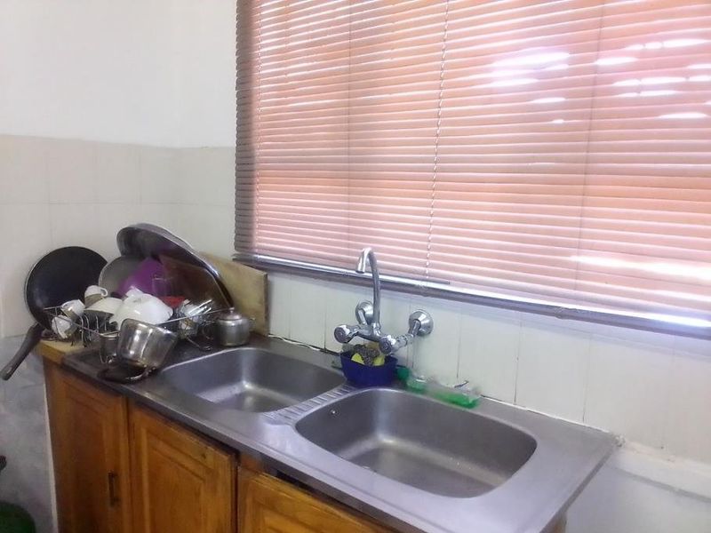 A dated kitchen area featuring a double stainless steel sink, wooden cabinetry, and tiled splashback, illuminated by natural light through venetian blinds.