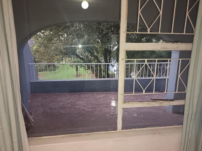 A covered balcony featuring brick paving, a metal security gate, and a metal railing overlooking a garden area.