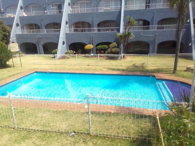 A communal swimming pool area featuring secure fencing and a well-maintained garden, set against the backdrop of a multi-story apartment building with private balconies.
