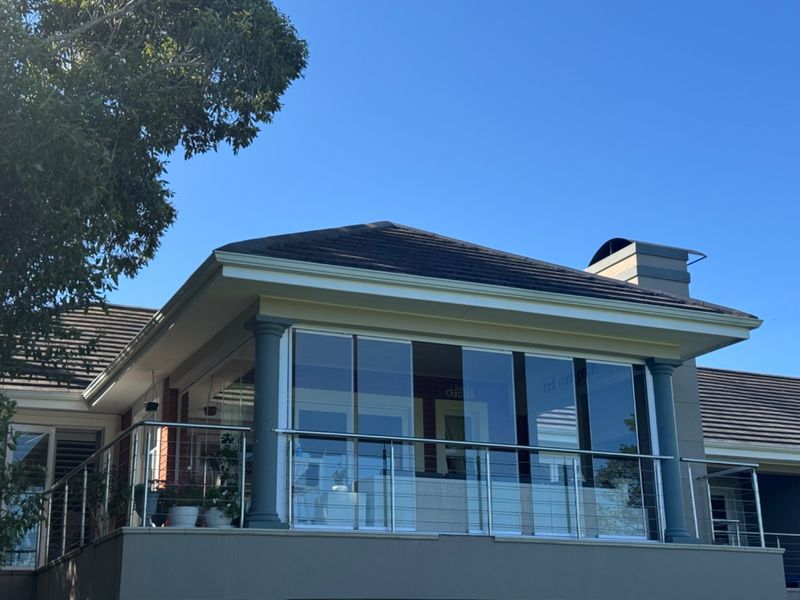 An elevated balcony featuring a modern glass balustrade with stainless steel railings, enclosed by sliding glass doors and sheltered by a tiled roof.