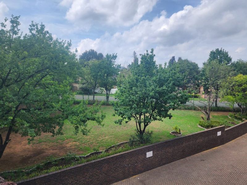 An elevated view overlooking a well-maintained communal garden area with mature trees, a brick boundary wall, and paved driveway access.