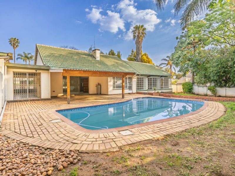 Rear exterior view featuring a swimming pool, covered patio area, and established garden with brick paving.