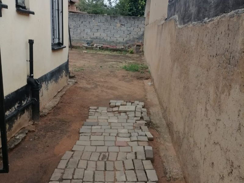 A narrow side yard area featuring a paved walkway, boundary wall, and exterior building wall with visible signs of wear and maintenance requirements.