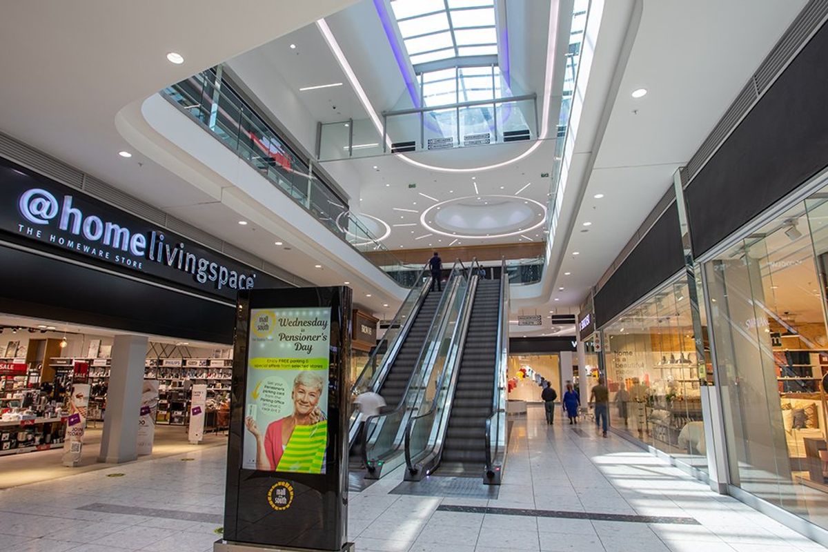 A modern commercial shopping mall atrium featuring a central escalator, expansive skylight, and multiple glass-fronted retail units with polished tile flooring.