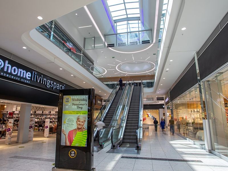 A modern commercial shopping mall atrium featuring a central escalator, expansive skylight, and multiple glass-fronted retail units with polished tile flooring.