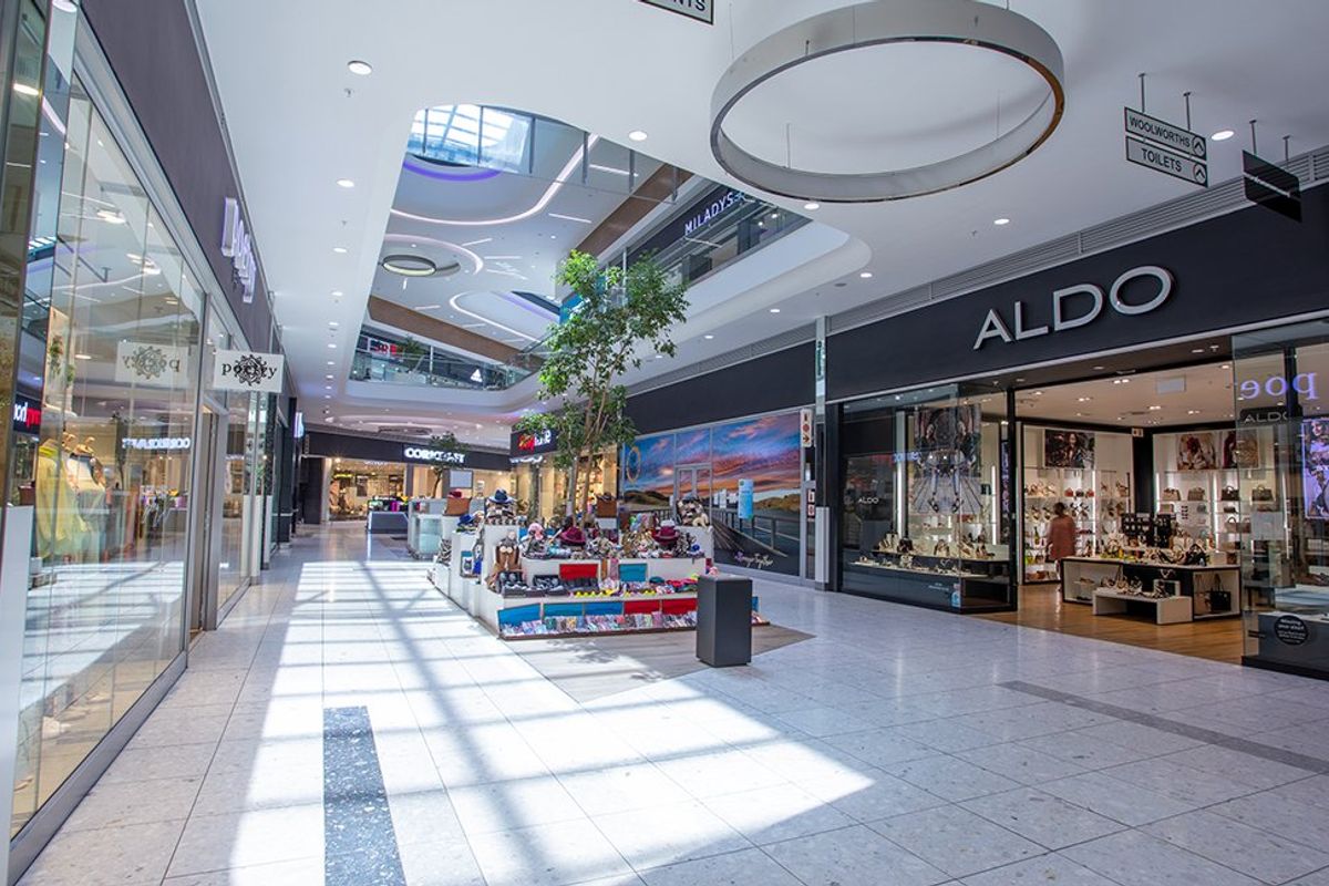 A bright, modern commercial shopping mall corridor featuring tiled floors, glass storefronts, and a large central skylight providing natural illumination.