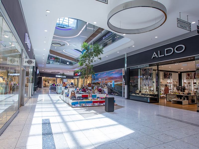 A bright, modern commercial shopping mall corridor featuring tiled floors, glass storefronts, and a large central skylight providing natural illumination.