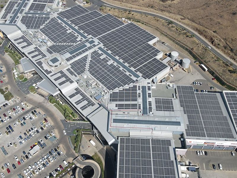 Aerial view of a large commercial shopping center featuring extensive rooftop solar panel installations, HVAC infrastructure, and surrounding asphalt parking facilities.