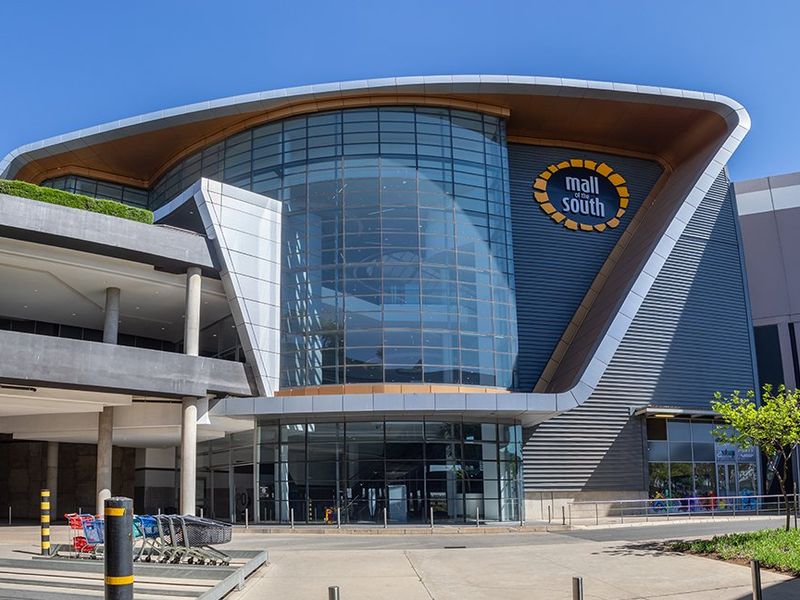 Modern commercial shopping center facade featuring a prominent glass curtain wall, metal architectural cladding, and a spacious covered entrance area.
