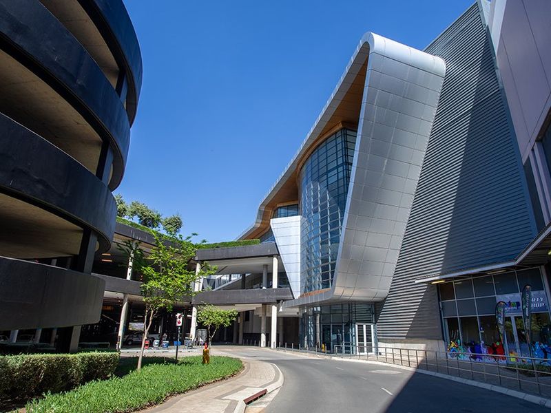 Modern commercial building exterior featuring contemporary metal cladding, extensive glass curtain walling, and an integrated multi-level parking structure with landscaped surroundings.