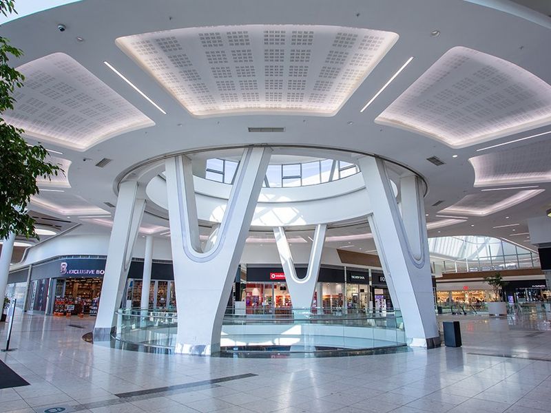 A modern, spacious commercial shopping mall atrium featuring high ceilings with recessed lighting, structural columns, and a central glass-enclosed void.