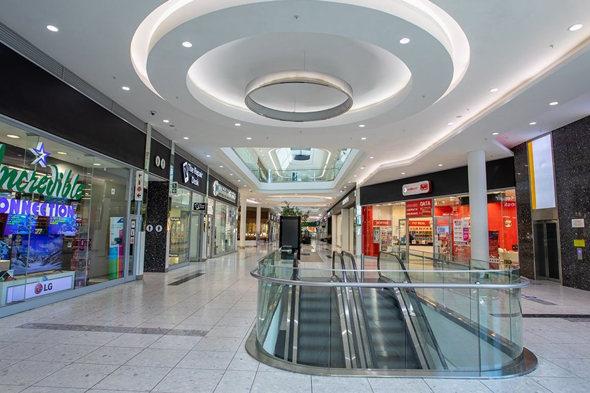 A modern commercial shopping mall corridor featuring high ceilings with recessed lighting, glass storefronts, and a central escalator access point.