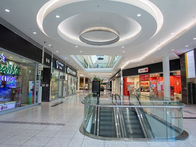 A modern commercial shopping mall corridor featuring high ceilings with recessed lighting, glass storefronts, and a central escalator access point.