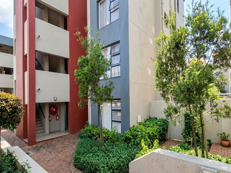 A well-maintained apartment complex exterior featuring a paved courtyard, landscaped garden areas, and accessible stairwell access.