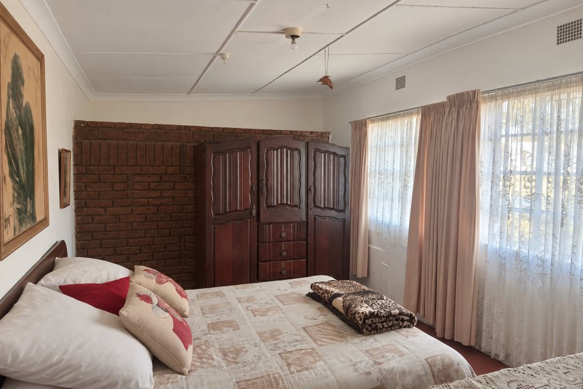 A bedroom featuring an exposed brick accent wall, tiled ceiling, and large windows with curtain rails, offering a dated but functional living space.