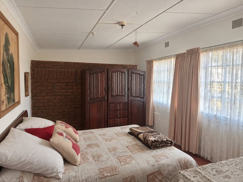 A bedroom featuring an exposed brick accent wall, tiled ceiling, and large windows with curtain rails, offering a dated but functional living space.