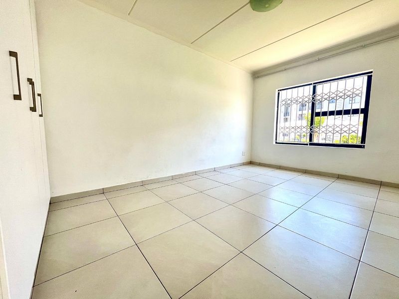 A bright, empty bedroom featuring neutral tiled flooring, built-in cupboards, and a window fitted with a security gate.
