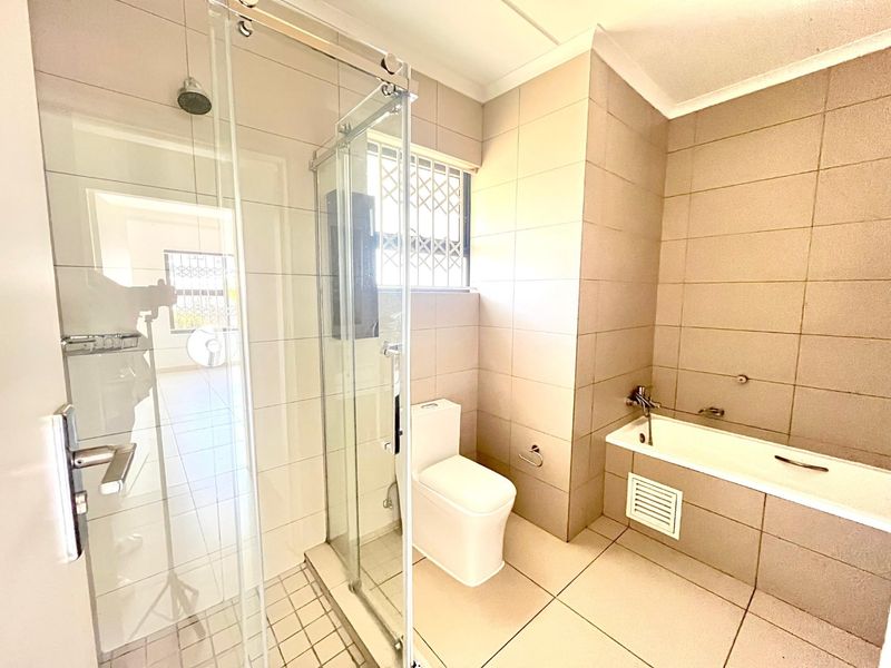 A clean, modern bathroom featuring a glass-enclosed shower, a built-in bathtub, and neutral floor-to-ceiling tiling with a secure window.