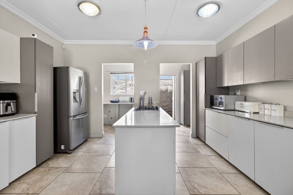 Modern kitchen featuring a central island with a gas cooktop, sleek cabinetry, stone countertops, and tiled flooring, leading to a scullery area.