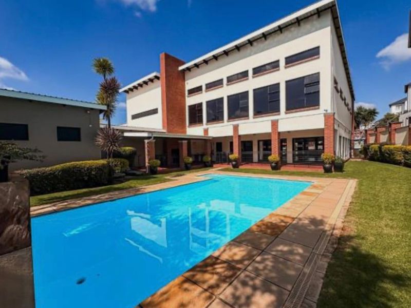 A well-maintained swimming pool area featuring a paved deck, landscaped garden, and a modern multi-story building facade with large windows.