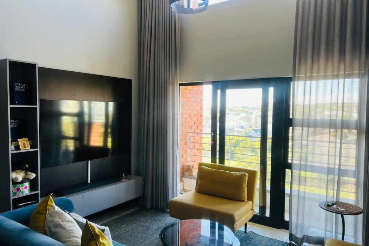 A bright, double-volume living room featuring a built-in media unit, floor-to-ceiling curtains, and direct access to a balcony with scenic views.